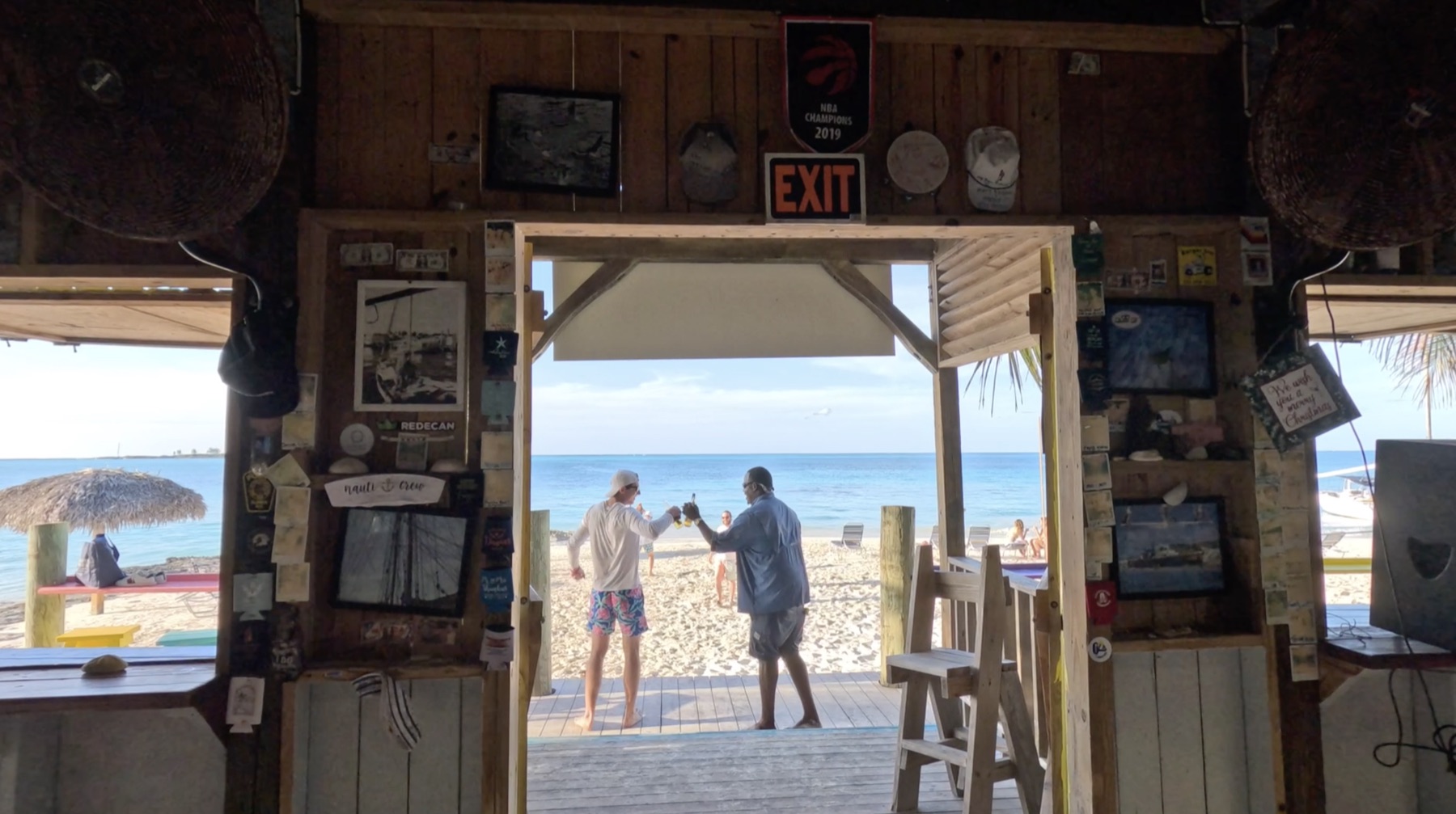 Two people touching bottles in the doorway of a beach bar hung with postcards, the sea beyond