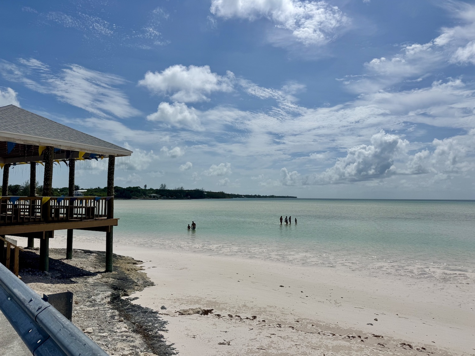 A covered beach bar on stilts at the edge of a white sand beach, with swimmers standing in calm shallow water