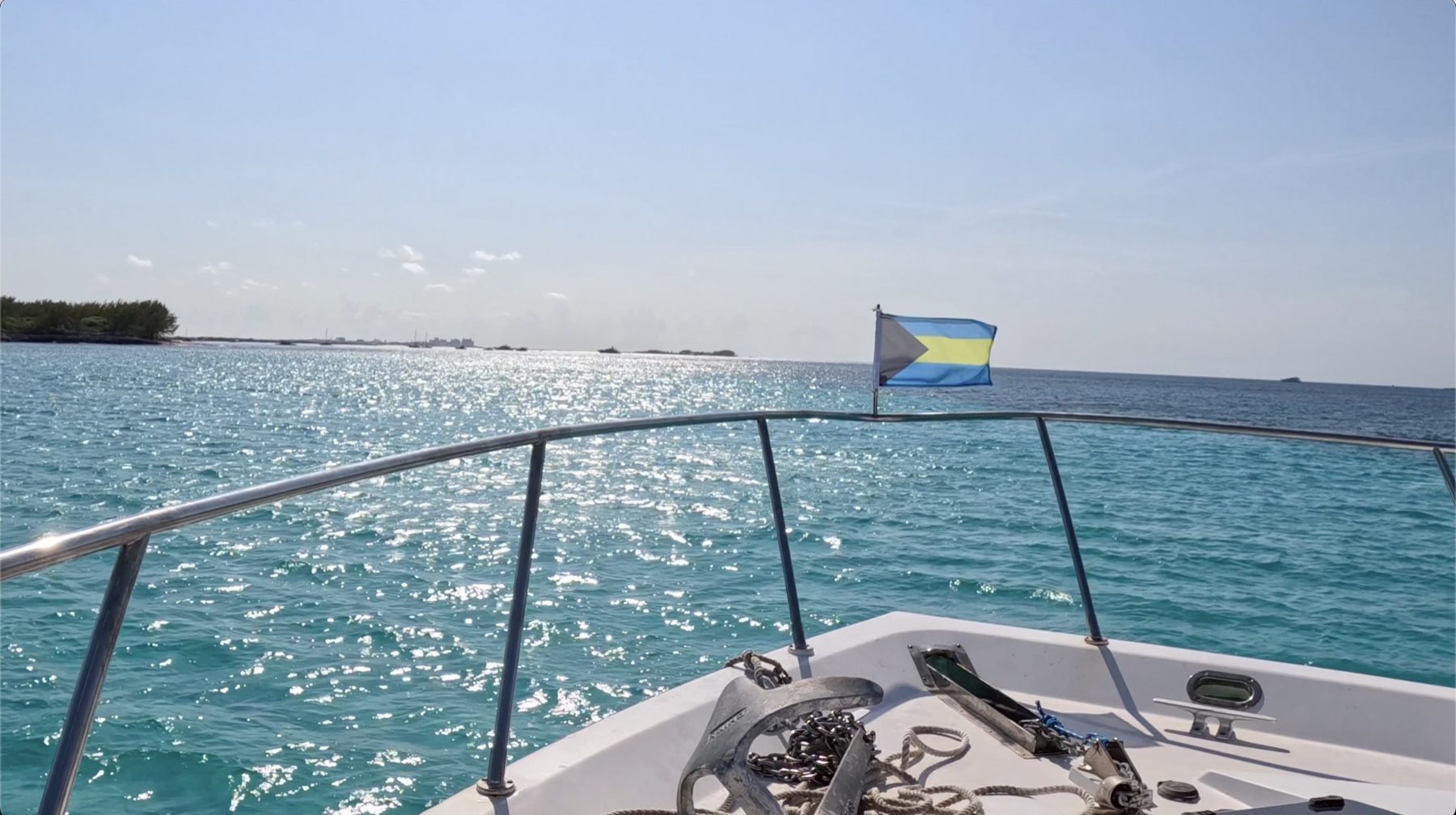 The bow of a boat under way in calm bright water, the Bahamian flag flying from the pulpit rail