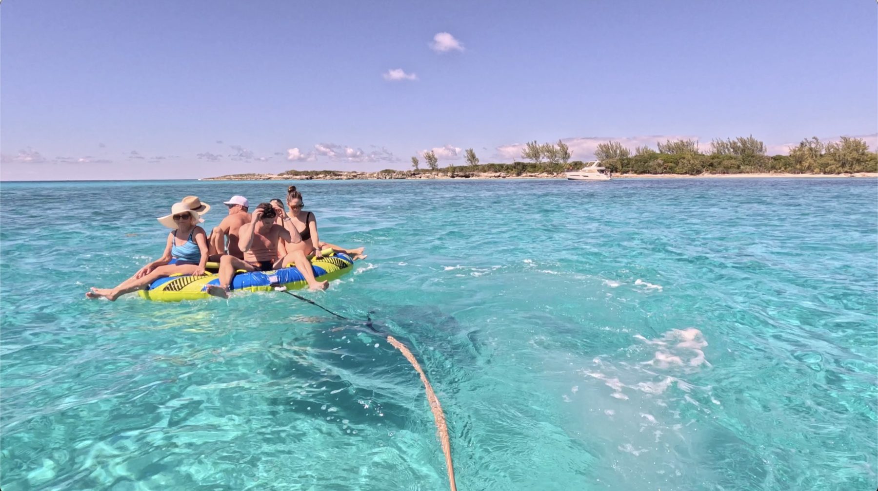 Four guests riding an inflatable towable across clear shallows, with a wooded cay and an anchored yacht beyond